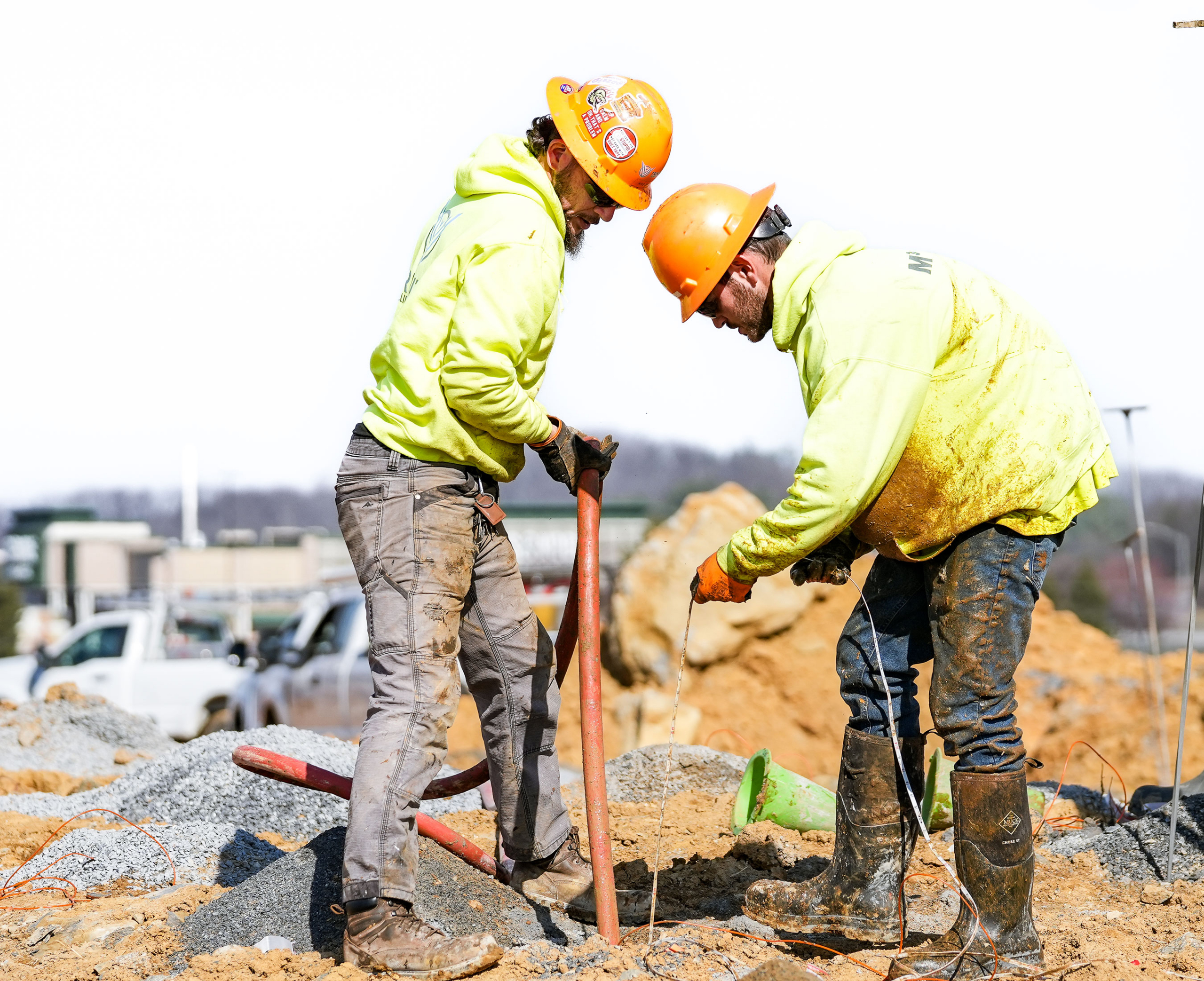 Two construction workers in bright yellow jackets and helmets focus on a task at a sandy site, wearing gloves and boots. The scene conveys teamwork.