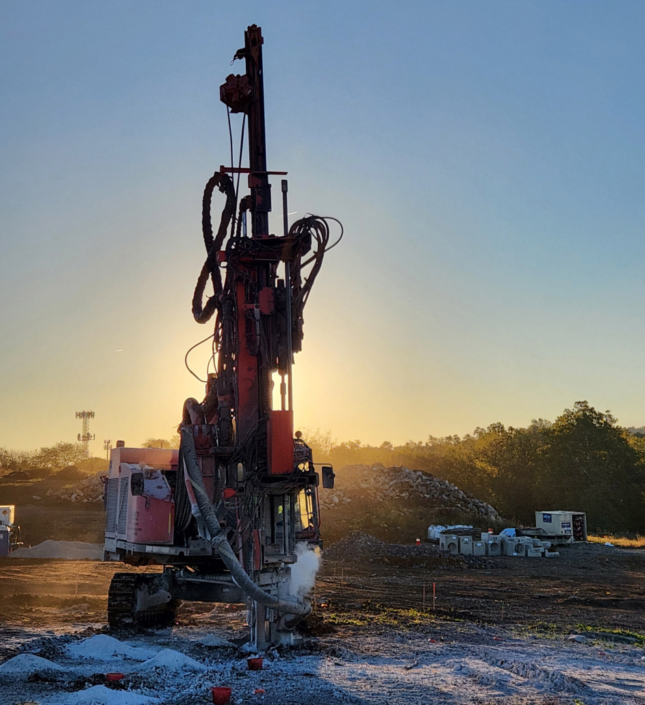 Trinity-Drilling-1 Sunrise over a construction site featuring a large red drilling machine in operation. The scene conveys industrious energy and early morning light.