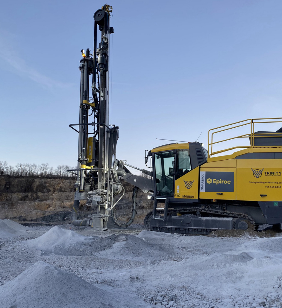 Trinity-Drilling-2 Yellow drilling machine in a quarry, labeled "Epiroc" and "Trinity." It stands on rocky terrain under a clear sky, conveying a sense of industrial strength.