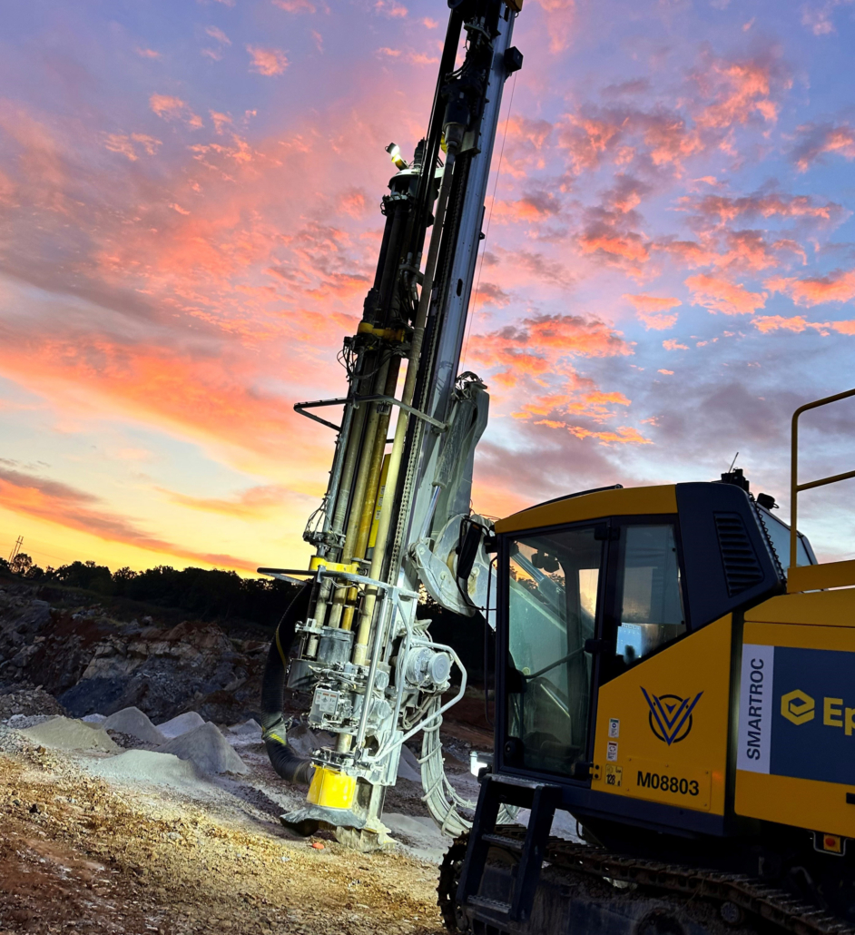 Trinity-Drilling-3 A large yellow drilling machine stands against a vibrant sunset, with hues of orange, pink, and purple clouds, creating a dramatic scene in an open quarry.