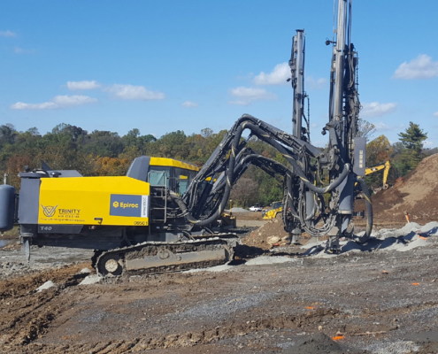 Trinity-Drilling-4 A large yellow Epiroc drill rig operates on a construction site, surrounded by dirt and machinery. Blue sky and trees are visible in the background.