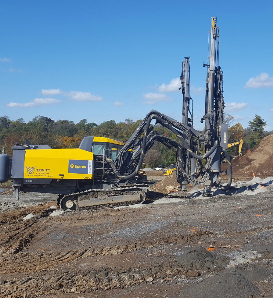 Trinity-Drilling-4 A large yellow Epiroc drill rig operates on a construction site, surrounded by dirt and machinery. Blue sky and trees are visible in the background.