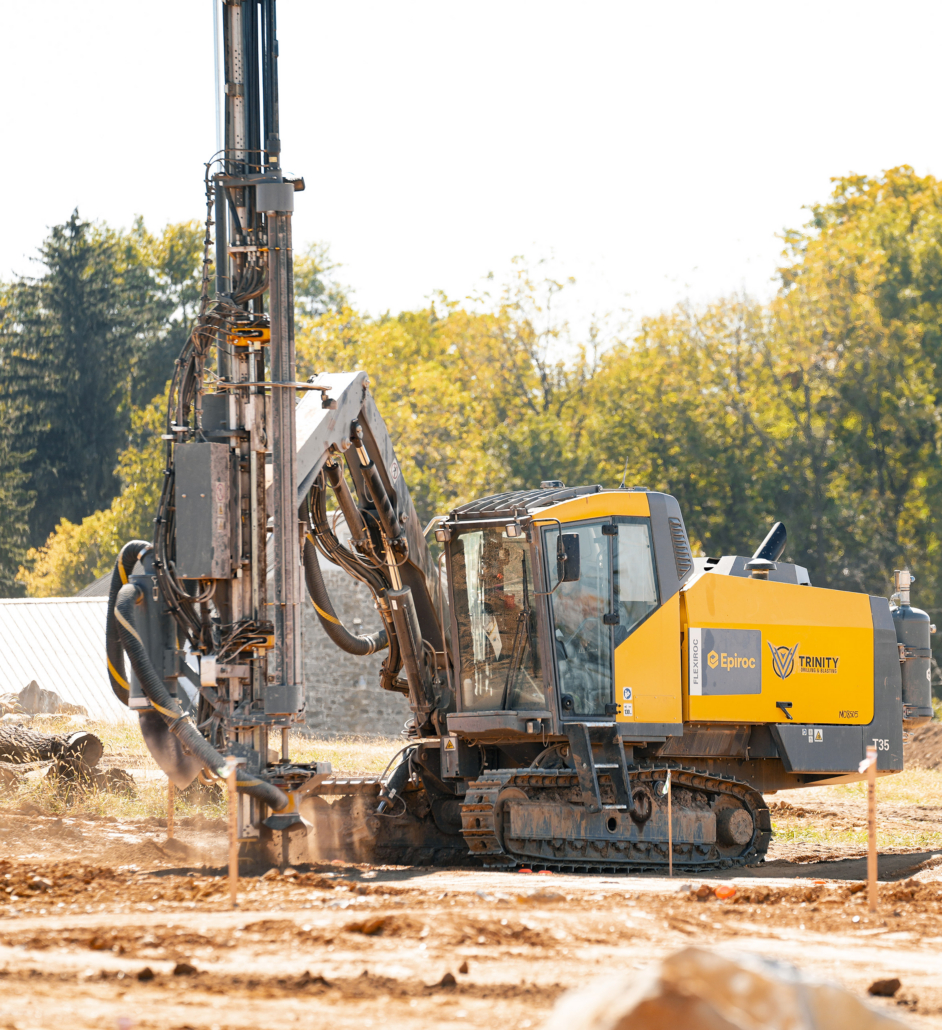 Trinity-Drilling-5 A yellow construction drilling machine operates on a dusty earth surface, surrounded by greenery and trees under a clear sky, conveying industriousness.