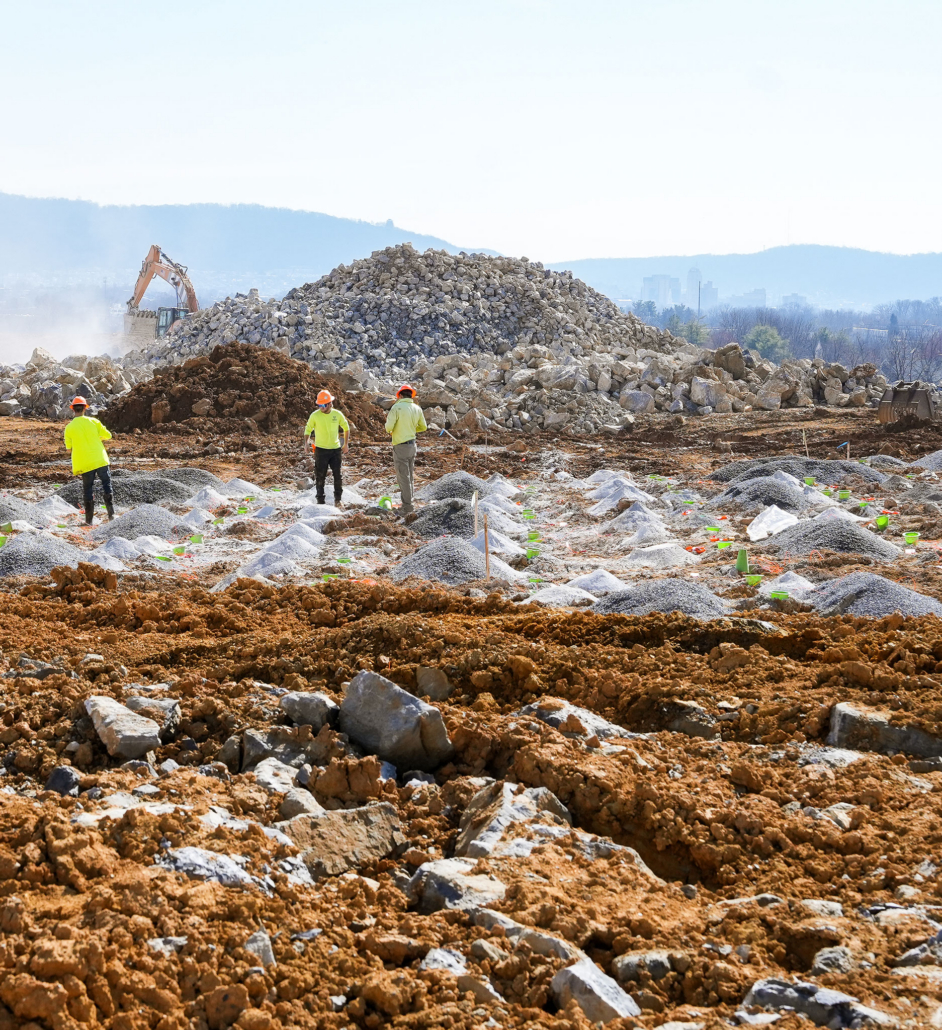 construction-infrastructure Construction site with three workers in yellow vests and helmets examining a rocky terrain. An excavator operates near a large pile of stones.