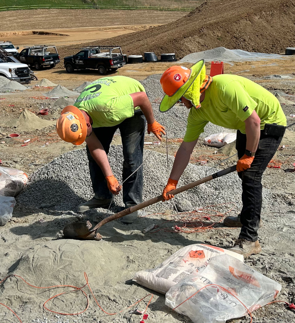 drilling-inset3 Two construction workers in bright green shirts and orange helmets are digging and using tools at a construction site. Trucks and materials are in the background.