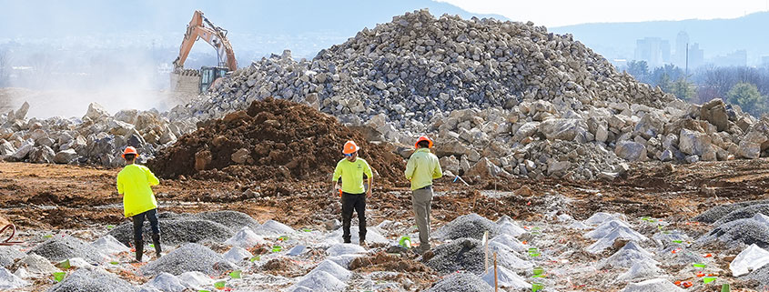 landscape_construction Construction site with workers in yellow vests and helmets examining a rocky terrain. An excavator operates near a large pile of stones.