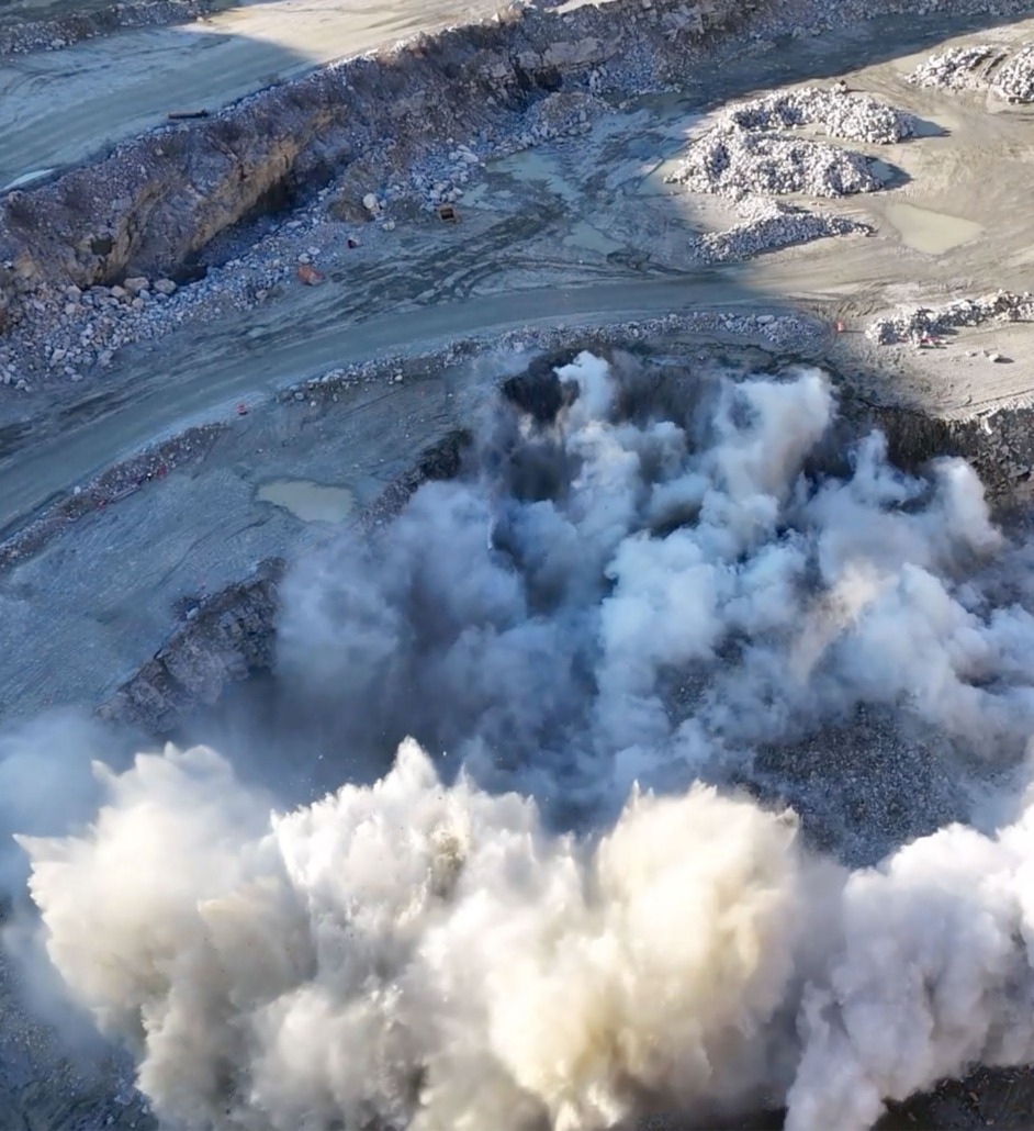 quarry-mining Aerial view of a quarry during a large explosion, with billowing white smoke clouds rising from the blast site. Surrounding rocky terrain appears rugged and barren.