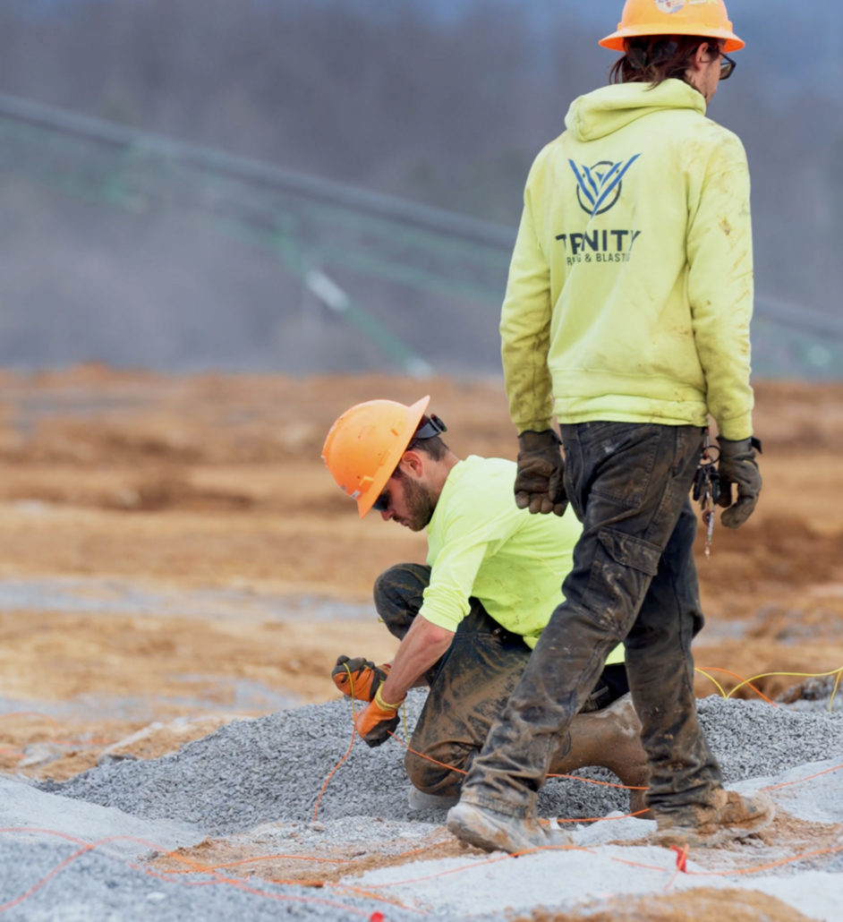 trinity-2-guys-onsite Two construction workers in safety gear are working on a gravel surface. One kneels, adjusting cables, while the other stands nearby. The mood is focused.