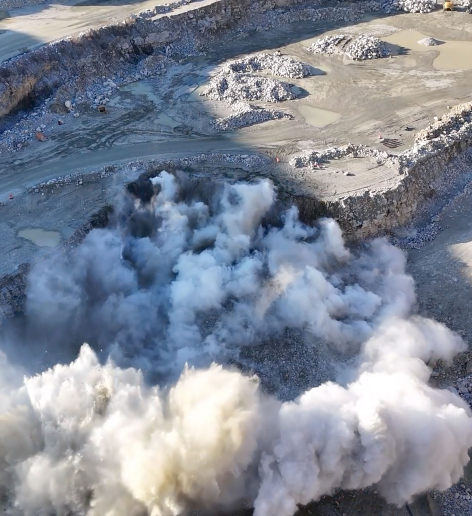 trinity-blasting-rocks1 Aerial view of a quarry during a large explosion, with billowing white smoke clouds rising from the blast site. Surrounding rocky terrain appears rugged and barren.