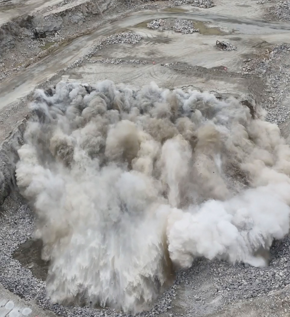 trinity-blasting-rocks2 Aerial view of a quarry during a large explosion, with billowing white smoke clouds rising from the blast site.