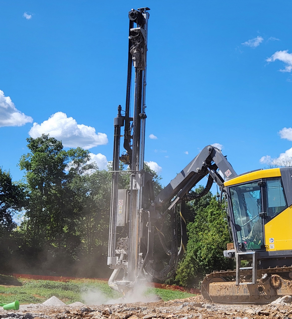 trinity-drilling Yellow construction vehicle with a tall drilling rig on a rocky site. Dust clouds billow as it operates. Green trees and blue sky in the background.