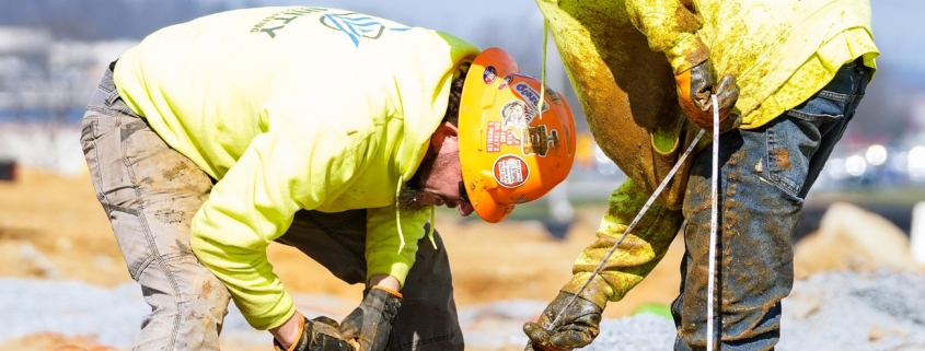 trinity-drilling1 Two construction workers in bright yellow jackets and hard hats work on a muddy site, handling tools and cables, with vehicles in the background.