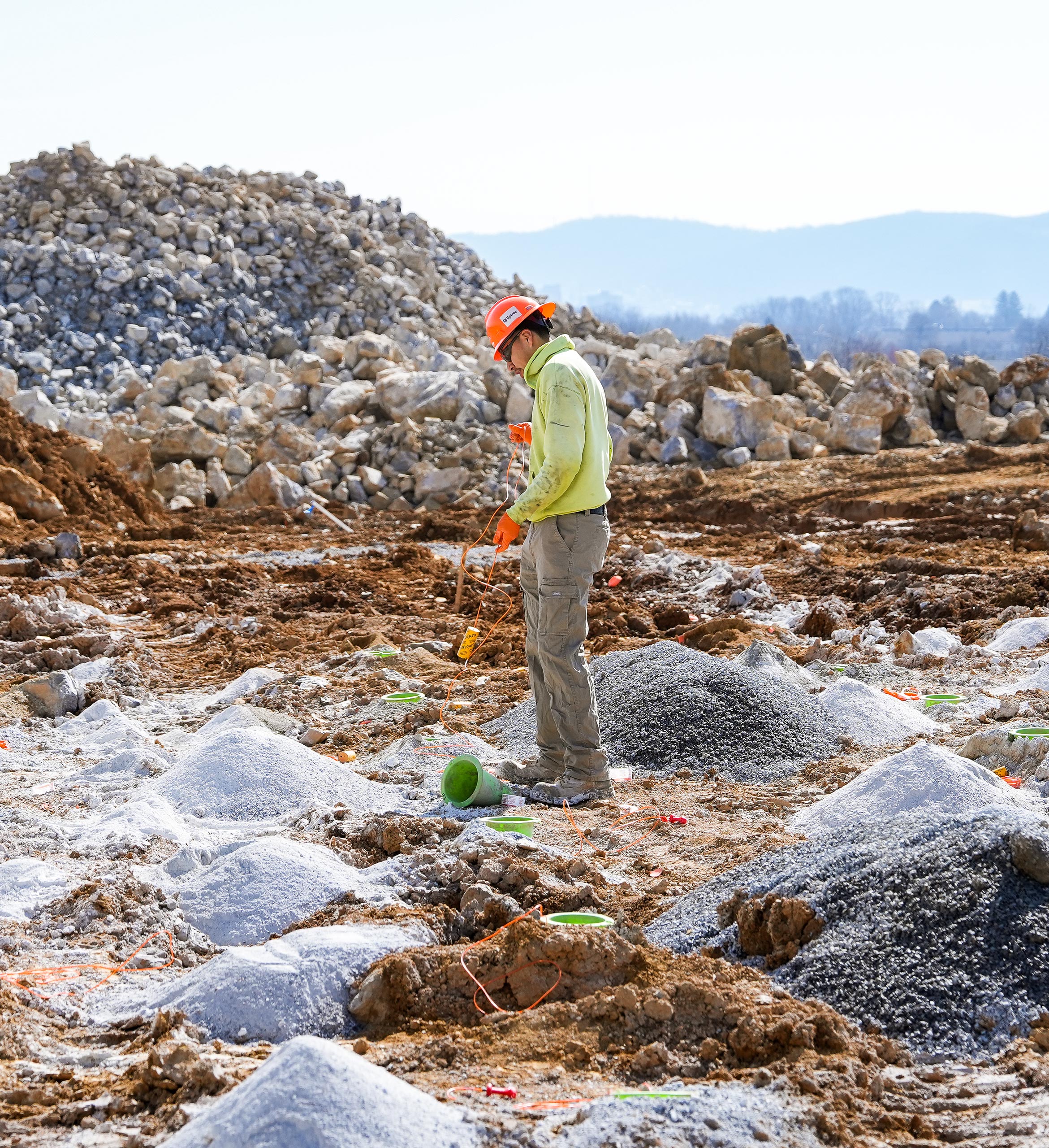 A construction worker wearing a hard hat and high-vis jacket stands on a rocky terrain, examining the ground. Piles of dirt and rocks surround him.
