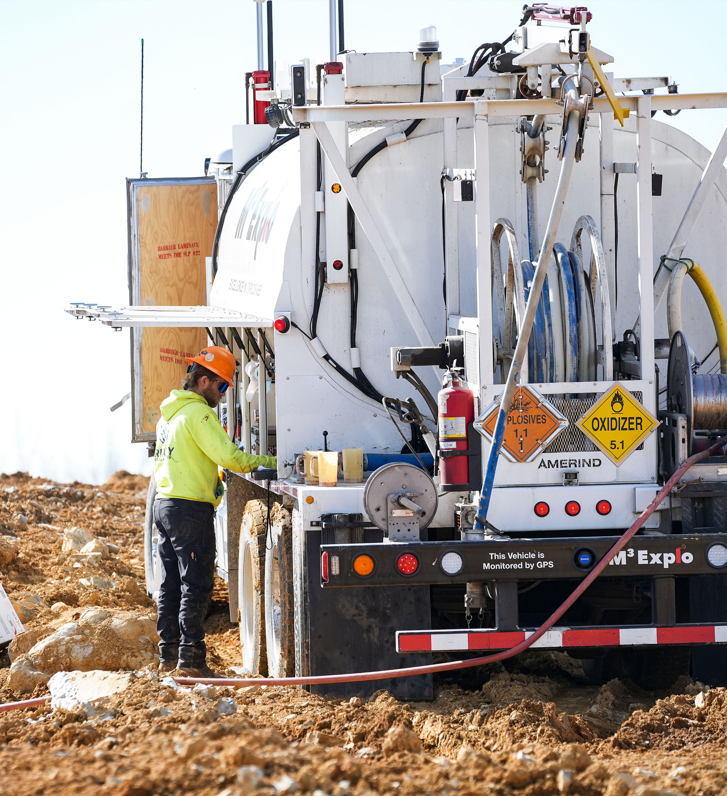 A worker in a yellow safety jacket and orange hard hat operates a large industrial vehicle on a dirt construction site.