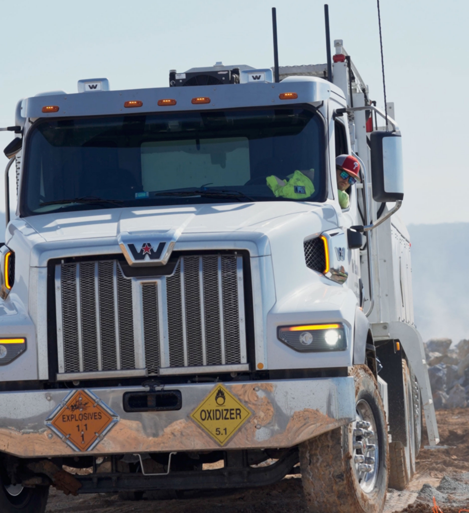 trinity-employee-driving-truck A white truck with prominent grille and hazard signs for explosives and oxidizers, driven by a person in a red cap, navigating a muddy terrain.