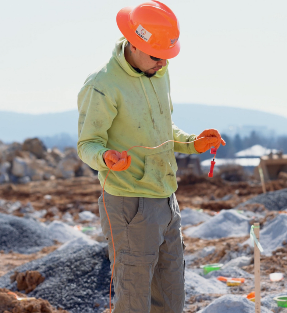 trinity-employee-with-detinator A construction worker in a bright orange hard hat and gloves inspects wiring at a rocky outdoor site, wearing a light green hoodie and gray pants.