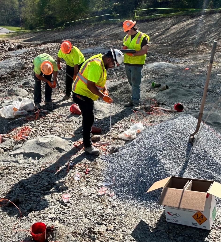 trinity-inset_4 Four construction workers in safety vests and helmets work on a gravel site. They handle wires near a pile of rock, suggesting preparation for blasting operations.