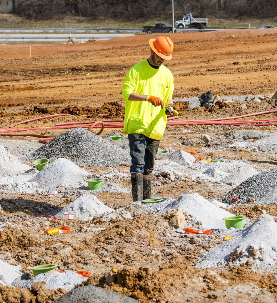 trinity-inset_4-edit A construction worker in a bright yellow shirt and orange hard hat examines soil at a worksite, surrounded by gravel mounds under a clear sky.