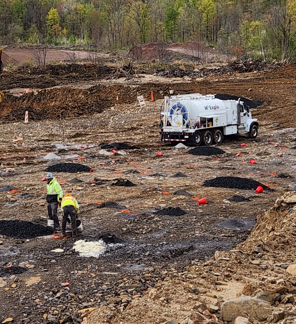 trinity-preblast Two workers in high-visibility jackets are inspecting a site with piles of earth, black material, and orange cones.