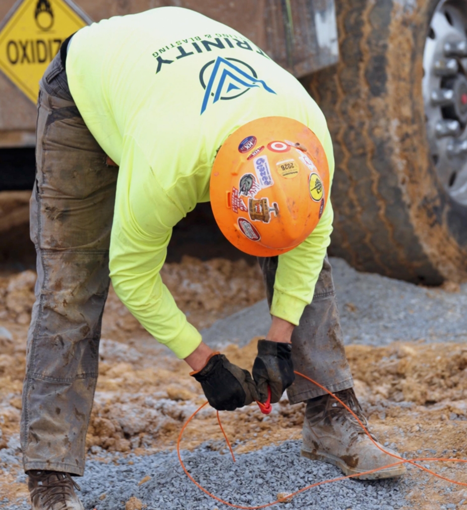 trinity_1 Construction worker in a neon shirt and orange hard hat crouches on gravel, handling orange cable. Background shows a muddy vehicle tire.