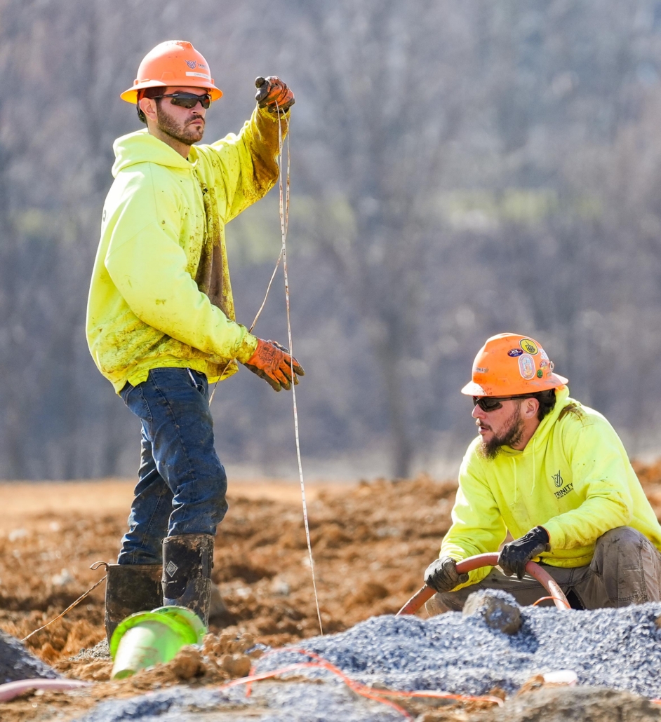 Two construction workers in bright yellow jackets and orange hard hats work on a rocky site. One stands holding a string, the other kneels, focusing intently.