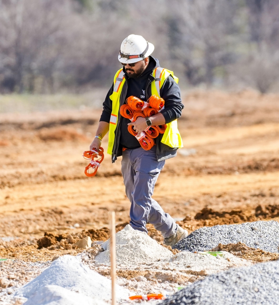 trinity_11 A construction worker in a yellow vest and helmet carries orange cables across a sandy construction site, conveying focus and industriousness.