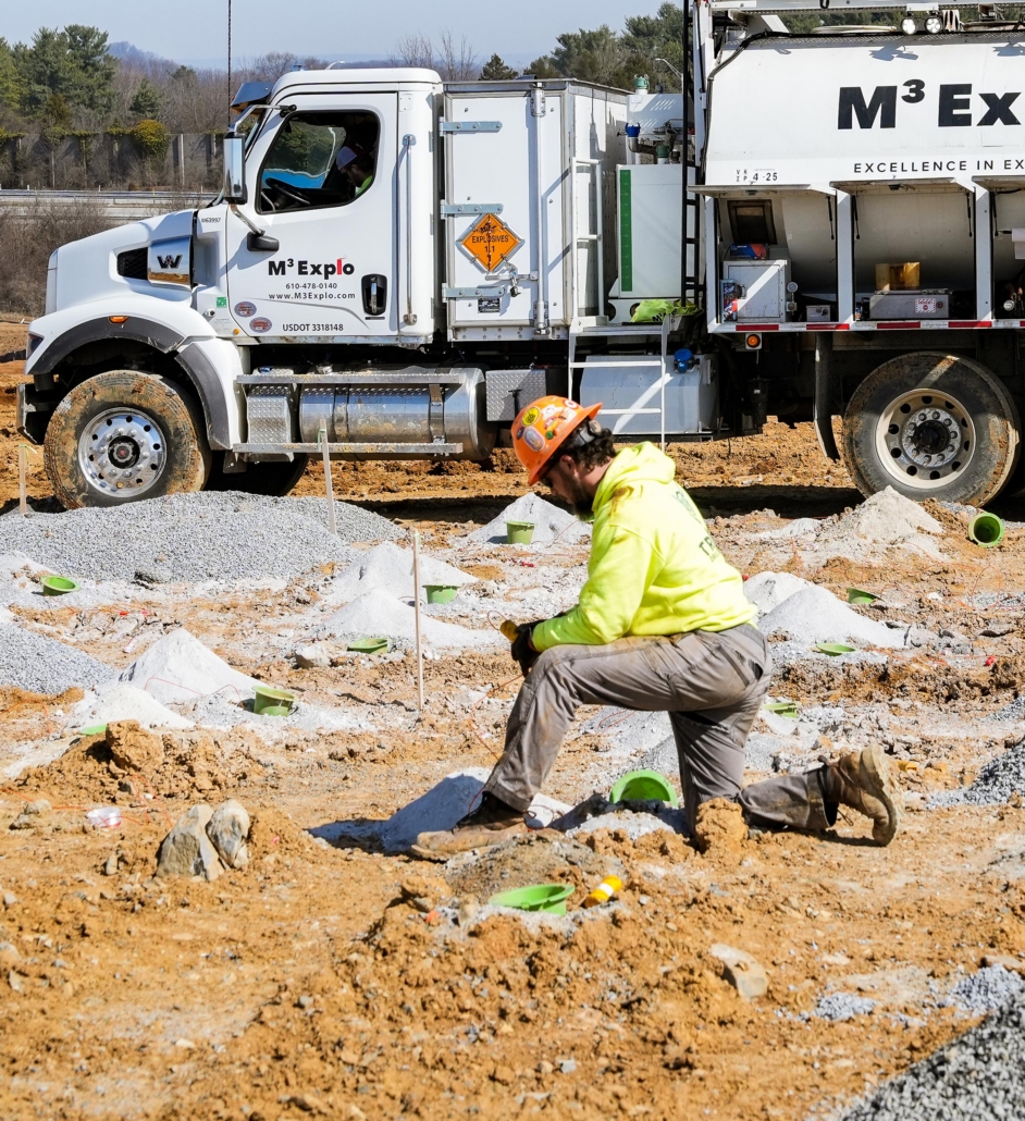 trinity_13 A construction worker in a neon jacket and hard hat kneels at a sandy site. A large M3 Explo truck and scattered equipment fill the background.