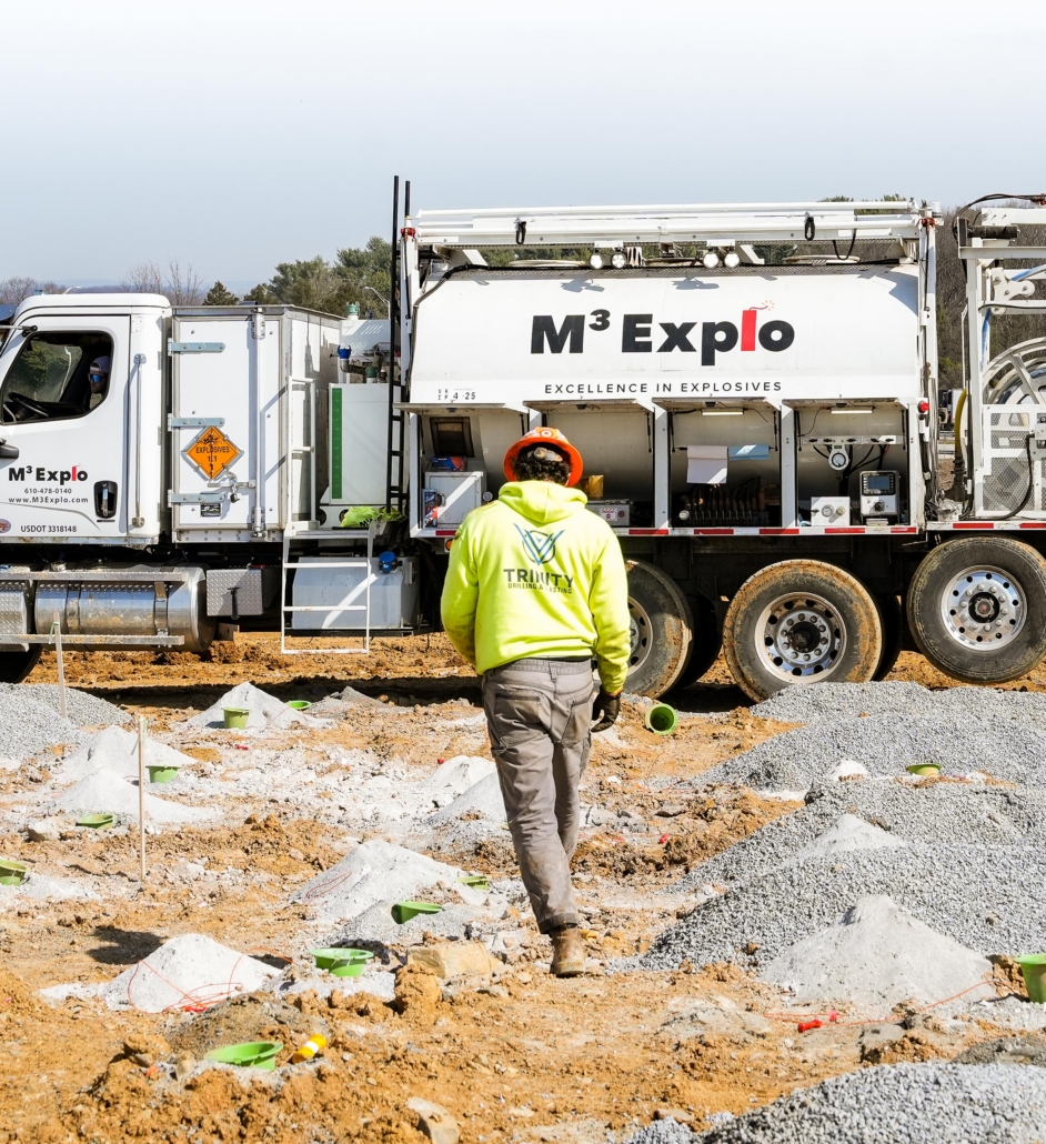 trinity_14 A construction worker in a neon yellow hoodie and hard hat walks towards an M3 Explo truck labeled "Excellence in Explosives" on a gravel site.
