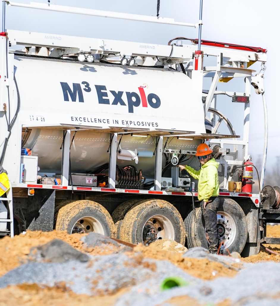 trinity_5 A worker in a bright green jacket and orange helmet operates equipment on a truck labeled "M³ Explo: Excellence in Explosives" at a construction site.