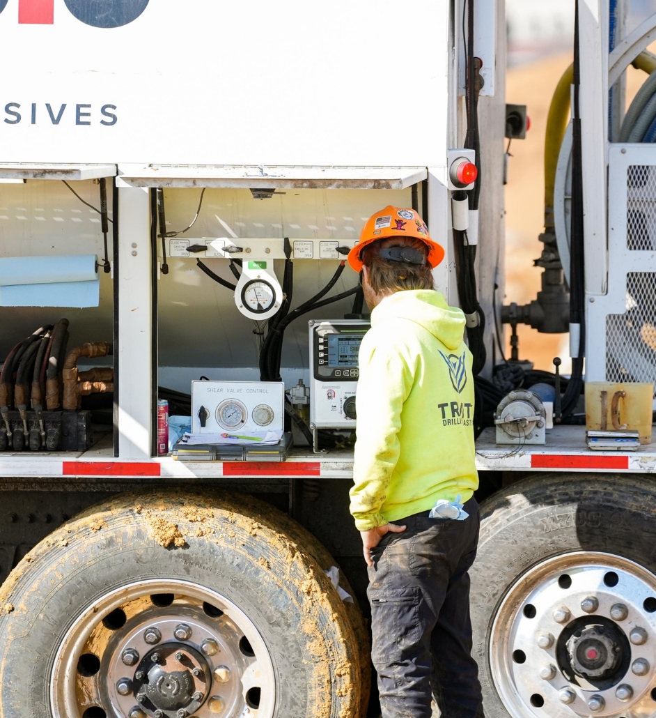 trinity_9 A construction worker in an orange hard hat and yellow hoodie stands by an equipment truck with gauges and controls. Mud adorns the truck's wheels.