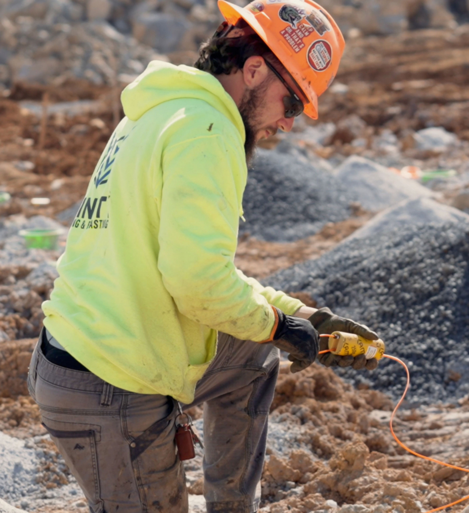 why-trinity_team-member-onsite Construction worker wearing a neon yellow hoodie and orange hard hat kneels on rocky ground, handling equipment with focus. The site is rough and active.